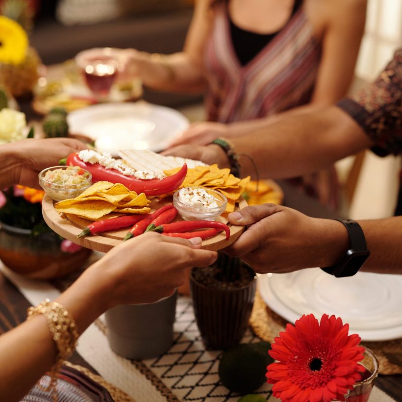 Hands of young woman passing tray with nachos, red hot chili peppers and savory sauce to male guest sitting by served festive table